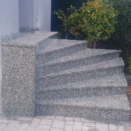 Spiralne schody zewnętrzne z szarego kamienia, prowadzące w górę obok zielonych krzewów i metalowej bramki.