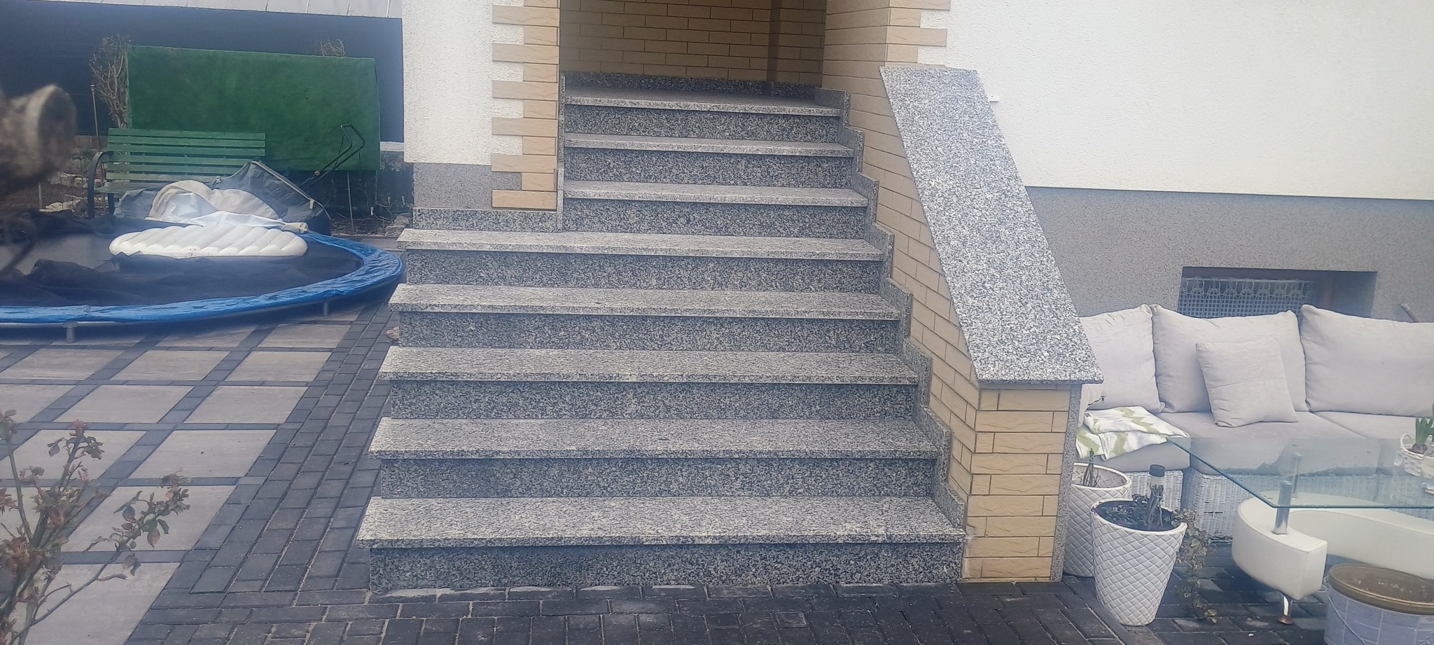 Zewnętrzne schody z szarego kamienia prowadzące do wejścia, z obudową z cegły klinkierowej, na tle kostki brukowej i mebli ogrodowych.
