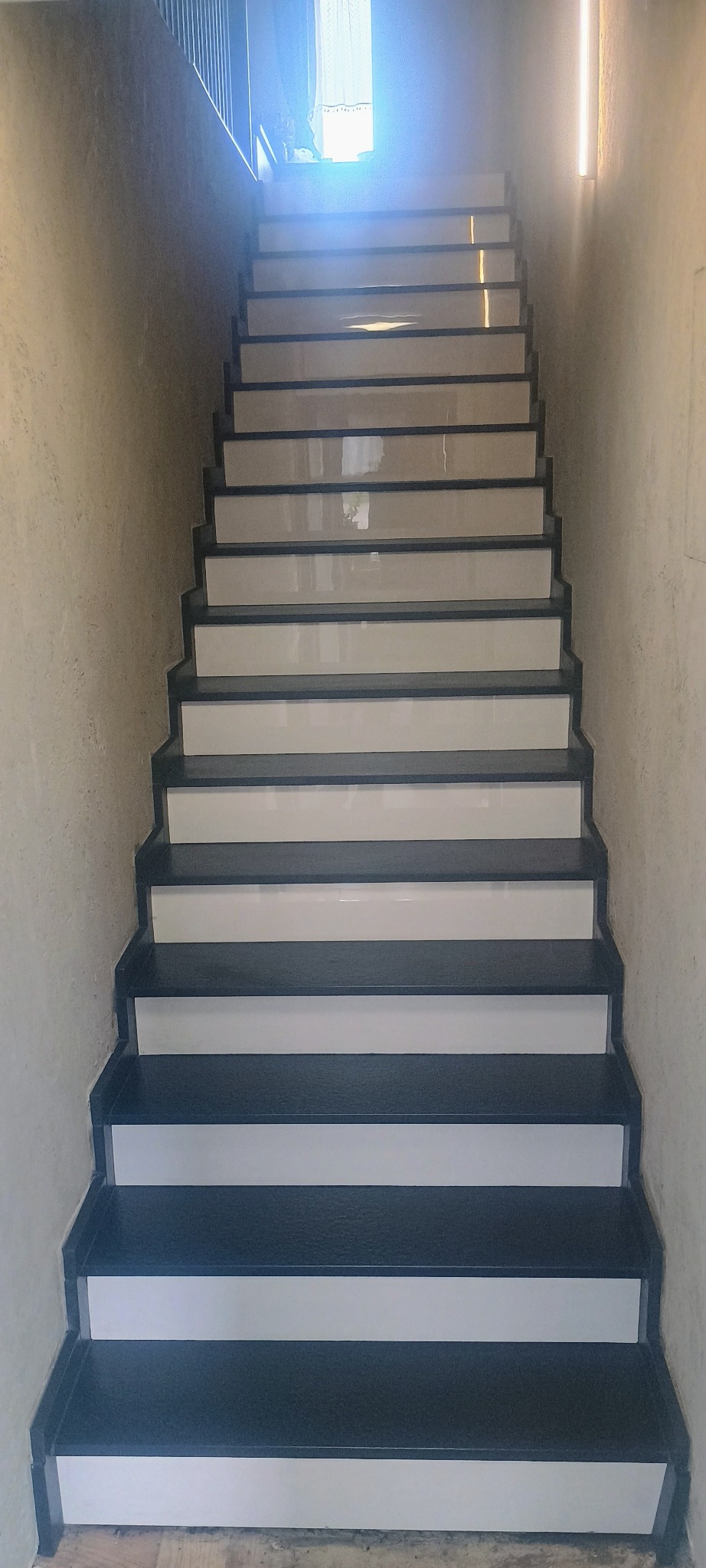 Wewnętrzne schody z ciemnego kamienia z białymi podstopnicami, widok z dołu do góry, z oświetleniem LED na ścianie po prawej stronie.