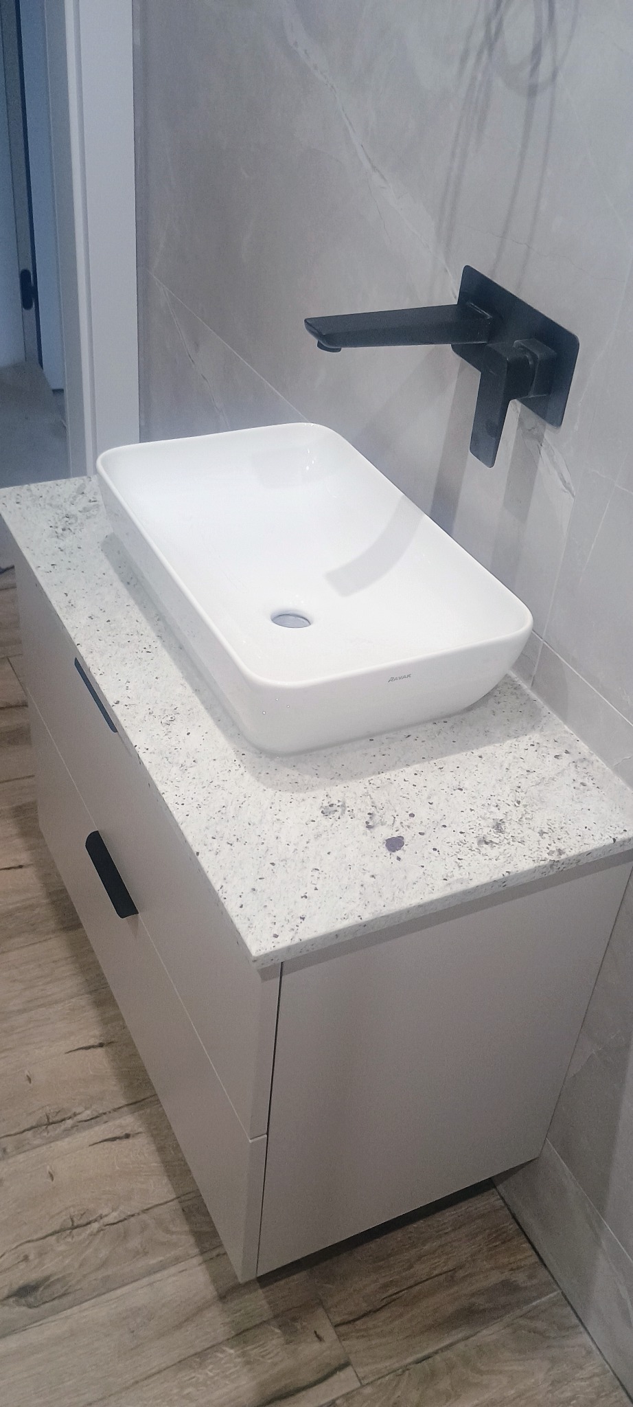Biała, prostokątna umywalka marki Ravak na blacie z jasnego kamienia z szarymi plamkami, zamontowana na szafce z szufladami; czarna, nowoczesna bateria ścienna, marmurowe płytki na ścianie w tle.