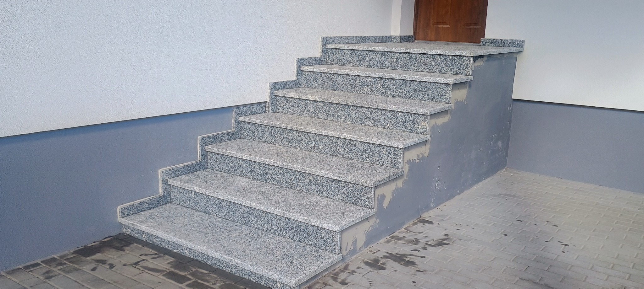 Zewnętrzne schody z szarego kamienia prowadzące do drzwi wejściowych, z widocznymi śladami poprawek tynkarskich na ścianie i podłodze.