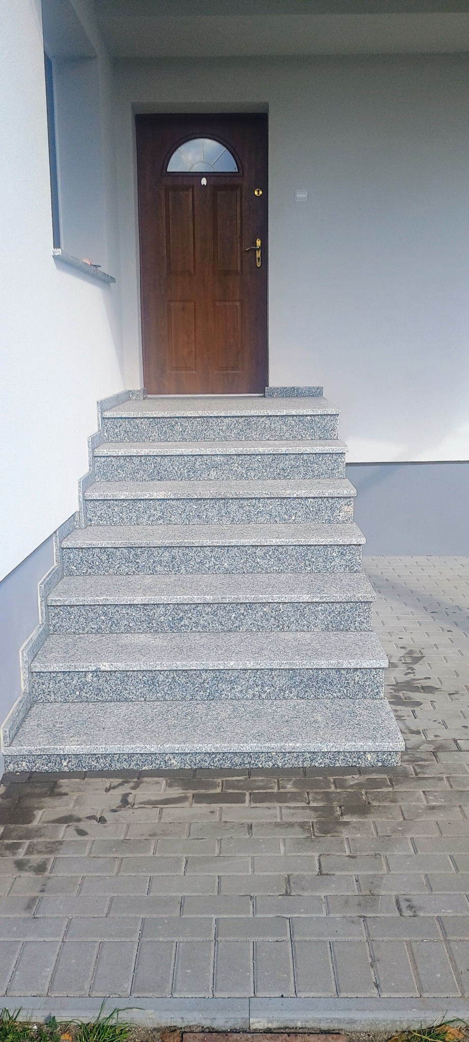 Zewnętrzne schody z szarego kamienia prowadzące do drewnianych drzwi wejściowych, z widoczną kostką brukową na podeście.