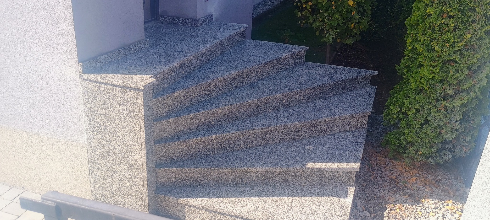 Zewnętrzne schody z szarego, nakrapianego kamienia prowadzące do wejścia budynku, z tujami w tle.
