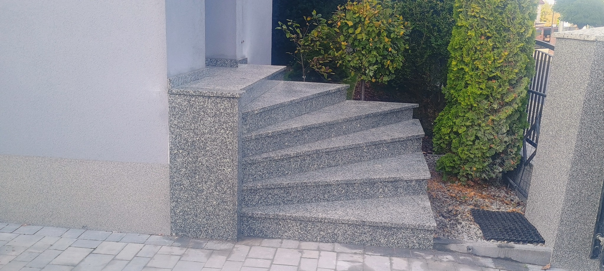 Spiralne schody zewnętrzne z szarego kamienia, prowadzące w górę obok zielonych krzewów i metalowej bramki.