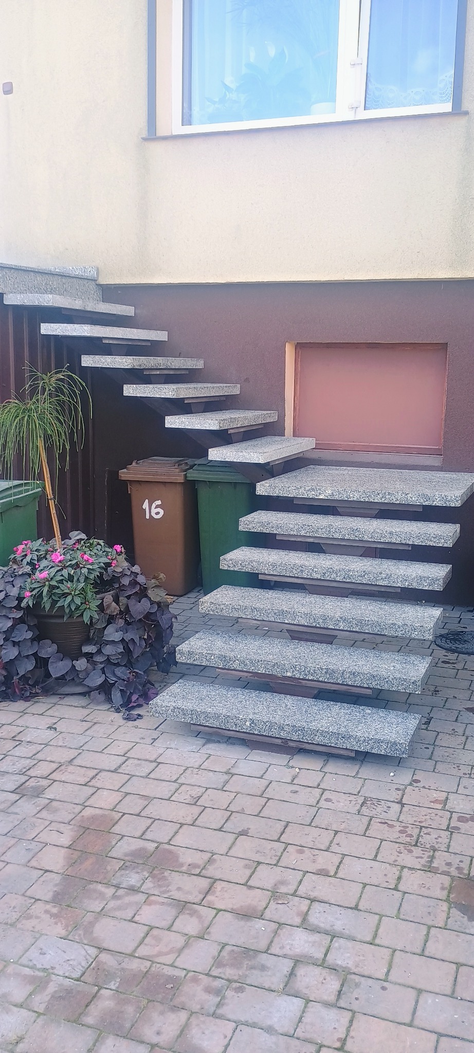 Zewnętrzne schody z szarego kamienia prowadzące do budynku, obok donica z kwiatami i kosze na śmieci.
