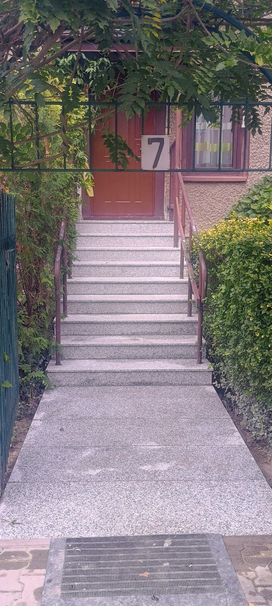 Granitowe schody wejściowe do domu z numerem 7, otoczone zielenią i metalowym ogrodzeniem.