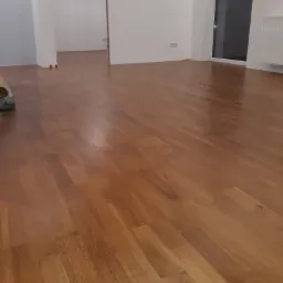 Wykończona podłoga z paneli drewnianych w pustym, jasnym pomieszczeniu z białymi ścianami; widoczny fragment opakowania paneli w lewym górnym rogu.