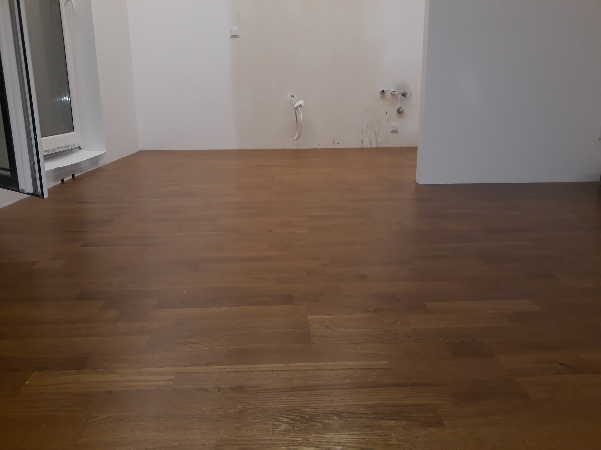 Położona, brązowa podłoga z paneli w pustym, jasnym pomieszczeniu z białymi ścianami i widocznymi instalacjami hydraulicznymi na ścianie.