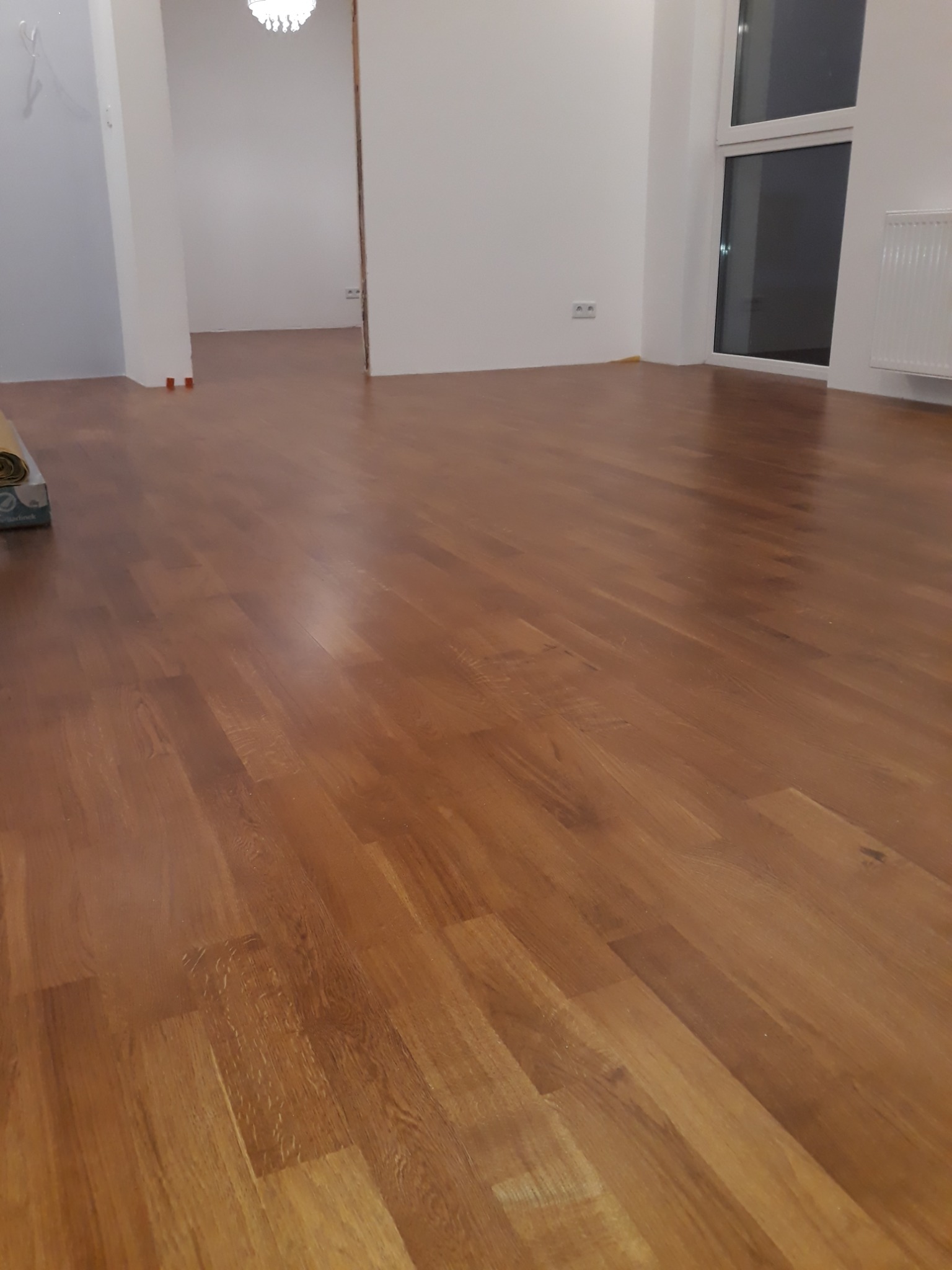 Wykończona podłoga z paneli drewnianych w pustym, jasnym pomieszczeniu z białymi ścianami; widoczny fragment opakowania paneli w lewym górnym rogu.