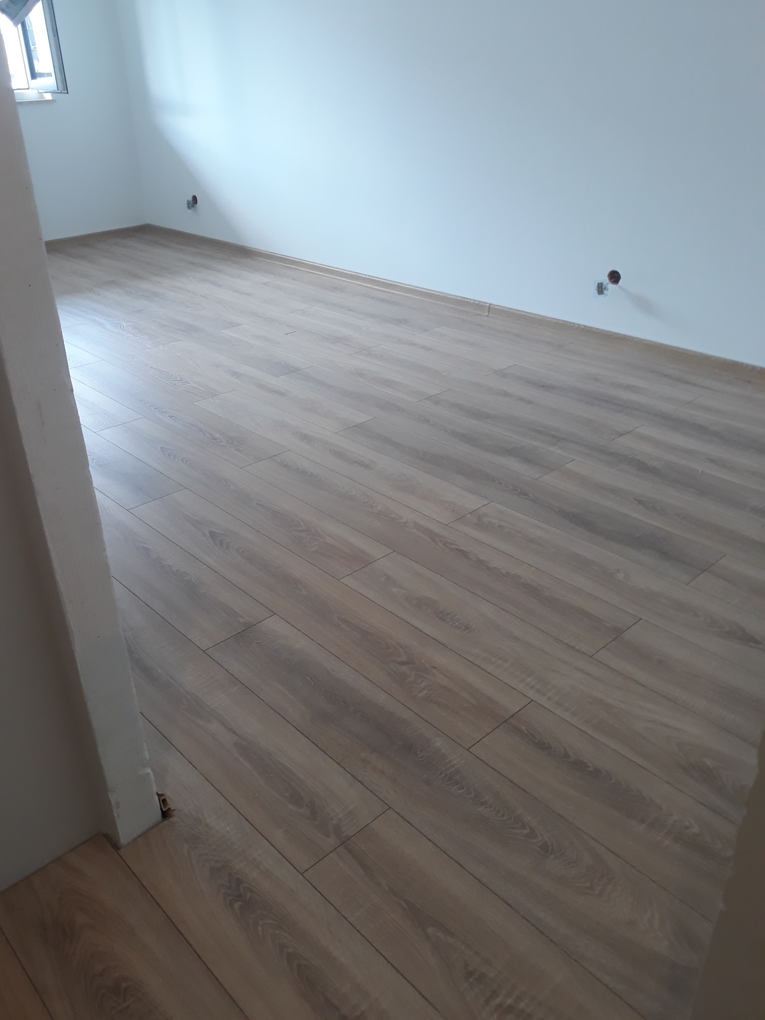 Nowo ułożone, jasnobrązowe panele podłogowe w pustym pokoju z białymi ścianami i wystającymi rurami instalacyjnymi.