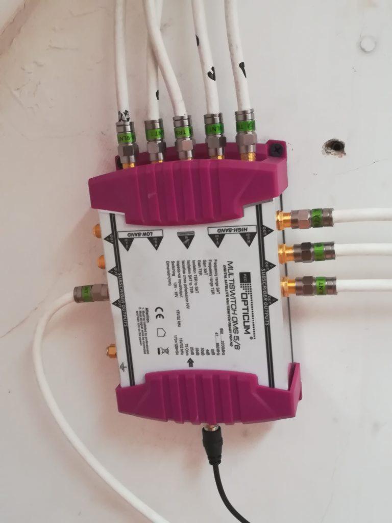 Rozdzielacz multiswitch Opticum OMS 5/6 zamontowany na białej ścianie, z podłączonymi białymi kablami koncentrycznymi, widoczne oznaczenia LOW-BAND i HIGH-BAND.