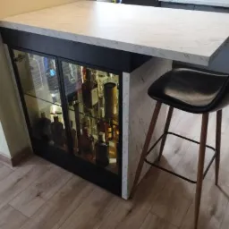 Wysoki blat kuchenny z marmurowym wzorem, wsparty na szafce z przeszklonymi drzwiami, w której widoczne są butelki alkoholu. Obok stoi czarny hoker z drewnianymi nogami.