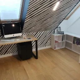 Pokój na poddaszu z biurkiem z jasnego drewna i czarnymi nogami, czarna komoda pod blatem, komputer iMac, ściana w czarno-białe pasy, białe regały na książki, drewniana podłoga i widok na zieloną...