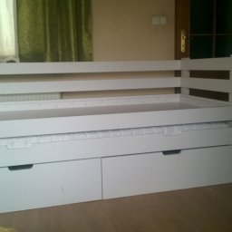 Mirosław Ciurej - Białe łóżko z szufladami na pościel, widoczne drewniane listwy konstrukcyjne i materac wsuwany pod spód, w tle okno z zasłonami i drzwi.