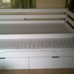 Mirosław Ciurej - Białe, drewniane łóżko piętrowe z wysuwanym dolnym łóżkiem i szufladami, bez pościeli, w pokoju z zasłoniętym oknem i ciemnymi drzwiami.