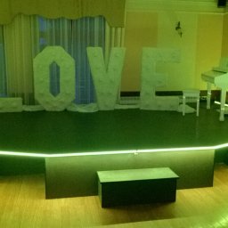 Mirosław Ciurej - Scena z napisem LOVE z dekoracyjnych liter, podświetlona zielonymi światłami, białe pianino na podeście, przygotowana na imprezę.