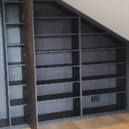 Zabudowa wnęki pod skosem z półkami, ciemne drewno, częściowo otwarte drzwiczki, widok na jasny parkiet i przeszklone drzwi.