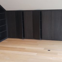 Mirosław Ciurej - Ciemnoszara zabudowa meblowa pod skosem, z szafkami z przesuwanymi drzwiami i otwartymi półkami, na jasnej drewnianej podłodze.