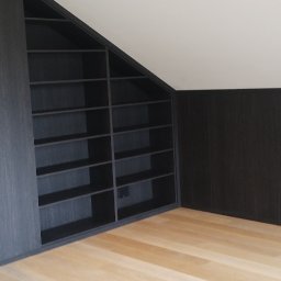 Mirosław Ciurej - Ciemna, drewniana szafa wnękowa z półkami, dopasowana do skosu dachu, z drabiną opartą o ścianę w tle.