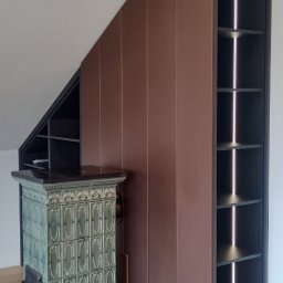 Mirosław Ciurej - Zabudowa wnęki na poddaszu z brązowymi, gładkimi frontami szafy, obok stoi piec kaflowy z zielonymi kaflami i czarnym blatem, a po prawej stronie otwarta, czarna półka z oświetleniem LED.