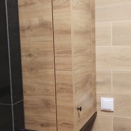 Mirosław Ciurej - Kuchenna szafka wisząca z frontami w kolorze jasnego drewna, zamontowana nad czarnym, błyszczącym blatem, z widocznym włącznikiem światła na ścianie z płytek.