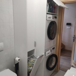 Mirosław Ciurej - Biała zabudowa w łazience z pralką i suszarką w kolumnie, otwarty kosz na pranie z kolorowymi ubraniami, widoczna toaleta i uchwyt na papier toaletowy.