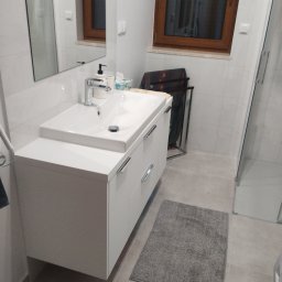 Mirosław Ciurej - Nowoczesna biała szafka łazienkowa z umywalką nablatową i chromowaną armaturą, szary dywanik, widok na okno z drewnianą ramą i kabinę prysznicową.