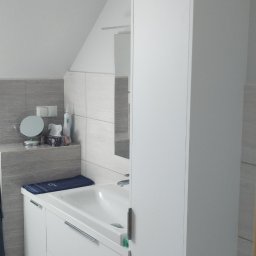 Mirosław Ciurej - Biała szafka łazienkowa z umywalką i wysoką szafką w nowoczesnej łazience z szarymi płytkami, widoczna część toalety.