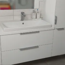 Nowoczesna, biała szafka łazienkowa z umywalką i dwoma szufladami, z wiszącą szafką boczną i toaletą w tle na tle jasnych płytek.