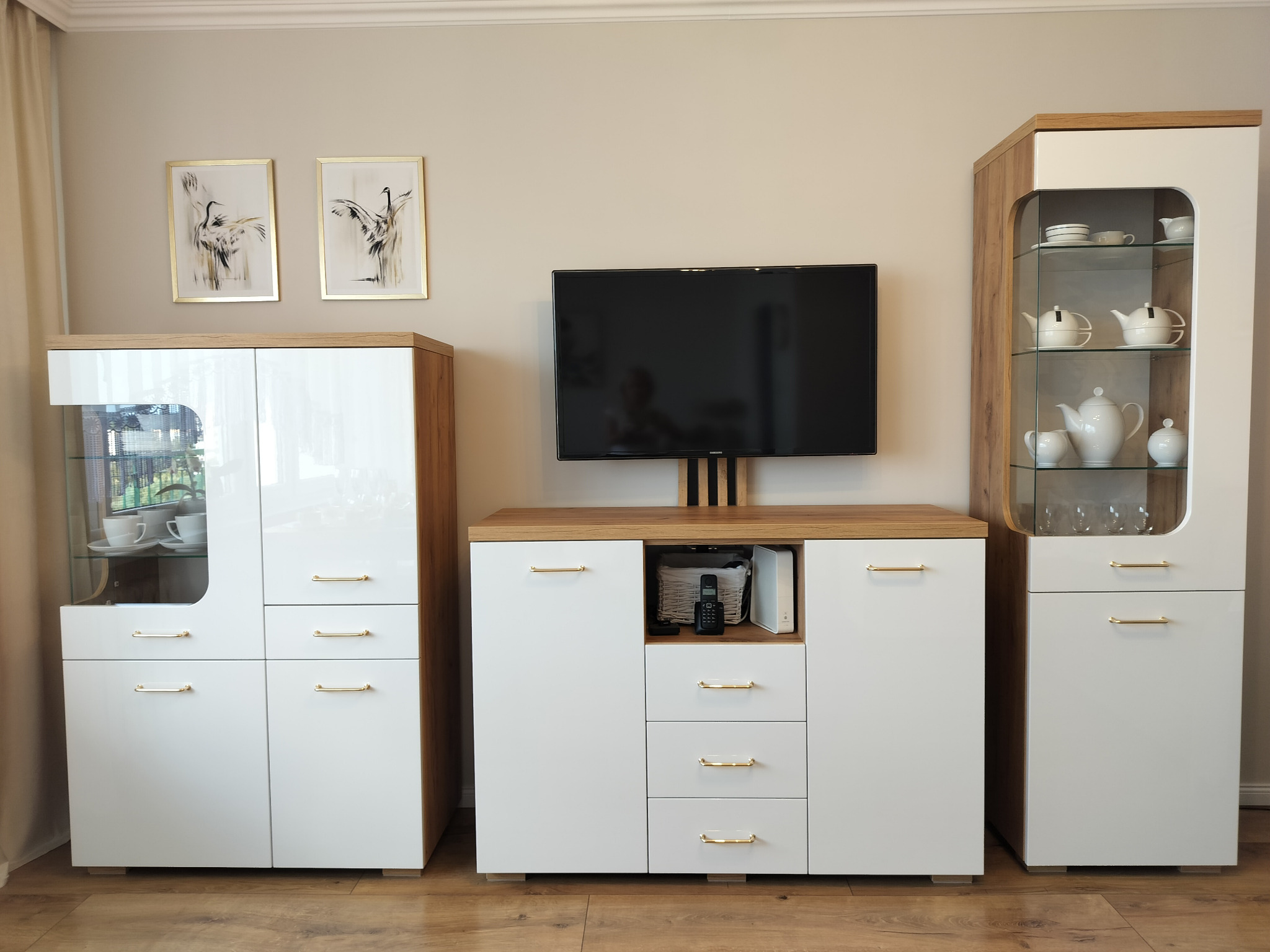 Nowoczesny zestaw mebli do salonu: białe fronty, drewnopodobne boki, witryny z ekspozycją porcelany, telewizor na komodzie, obrazy z motywem żurawi.