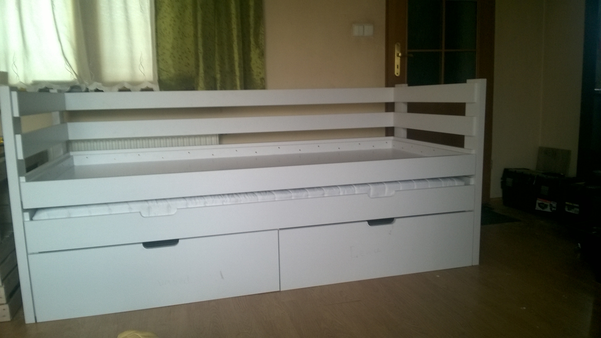 Białe łóżko z szufladami na pościel, widoczne drewniane listwy konstrukcyjne i materac wsuwany pod spód, w tle okno z zasłonami i drzwi.