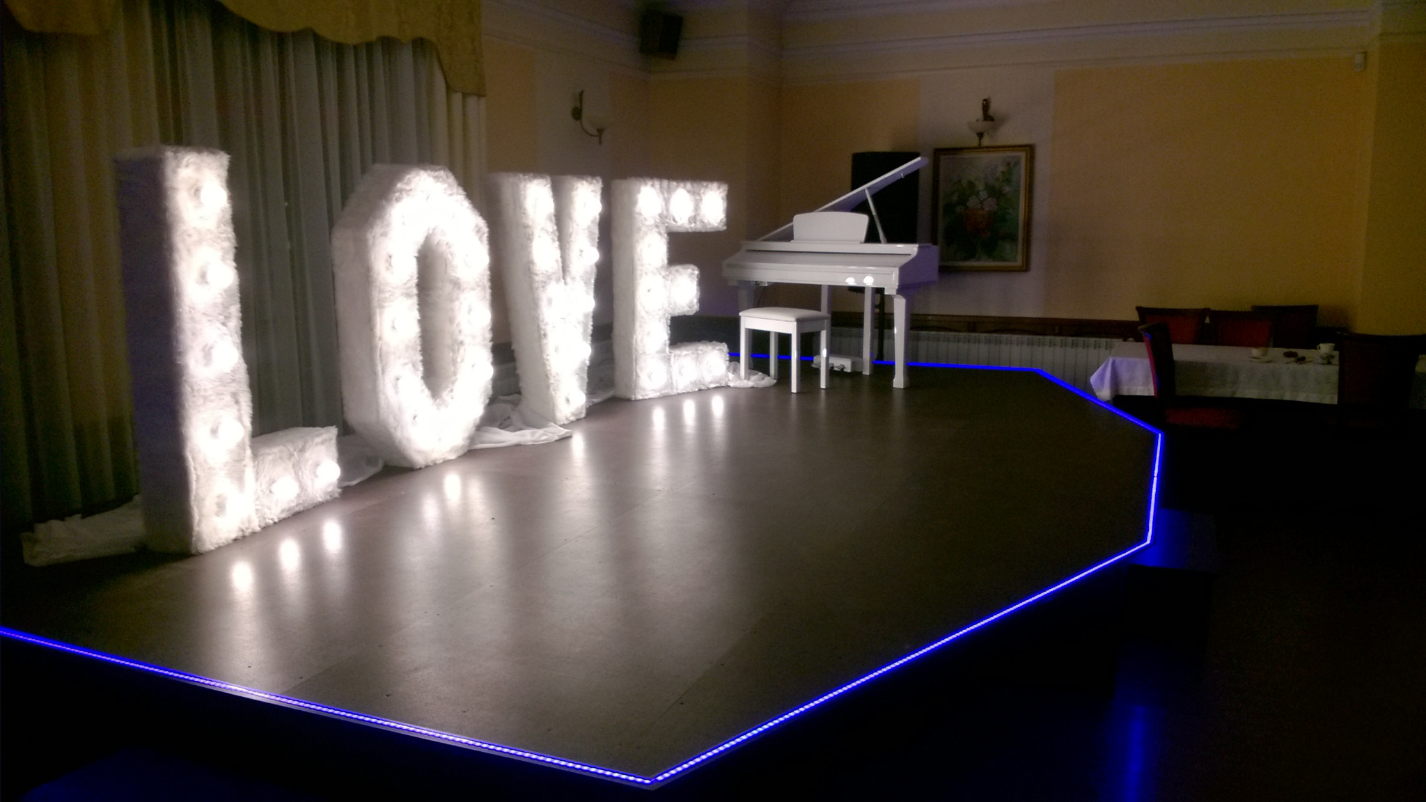 Scena z napisem LOVE wykonanym z podświetlanych liter obitych futerkiem, białe pianino w tle, niebieskie oświetlenie krawędzi sceny, sala bankietowa.