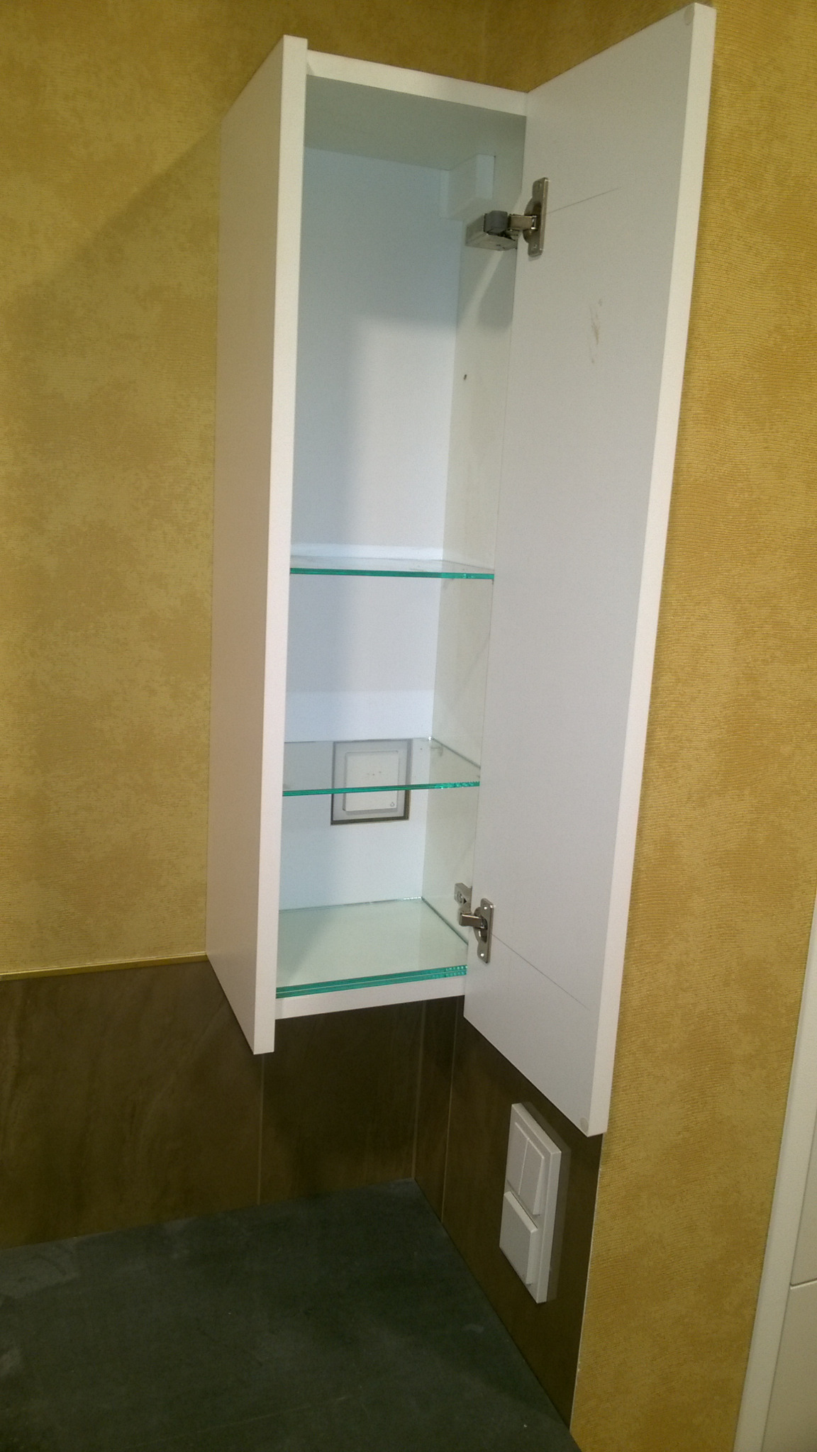 Biała szafka wisząca z otwartymi drzwiczkami, szklane półki wewnątrz, widoczne okucia, żółta ściana w tle, ciemna podłoga.
