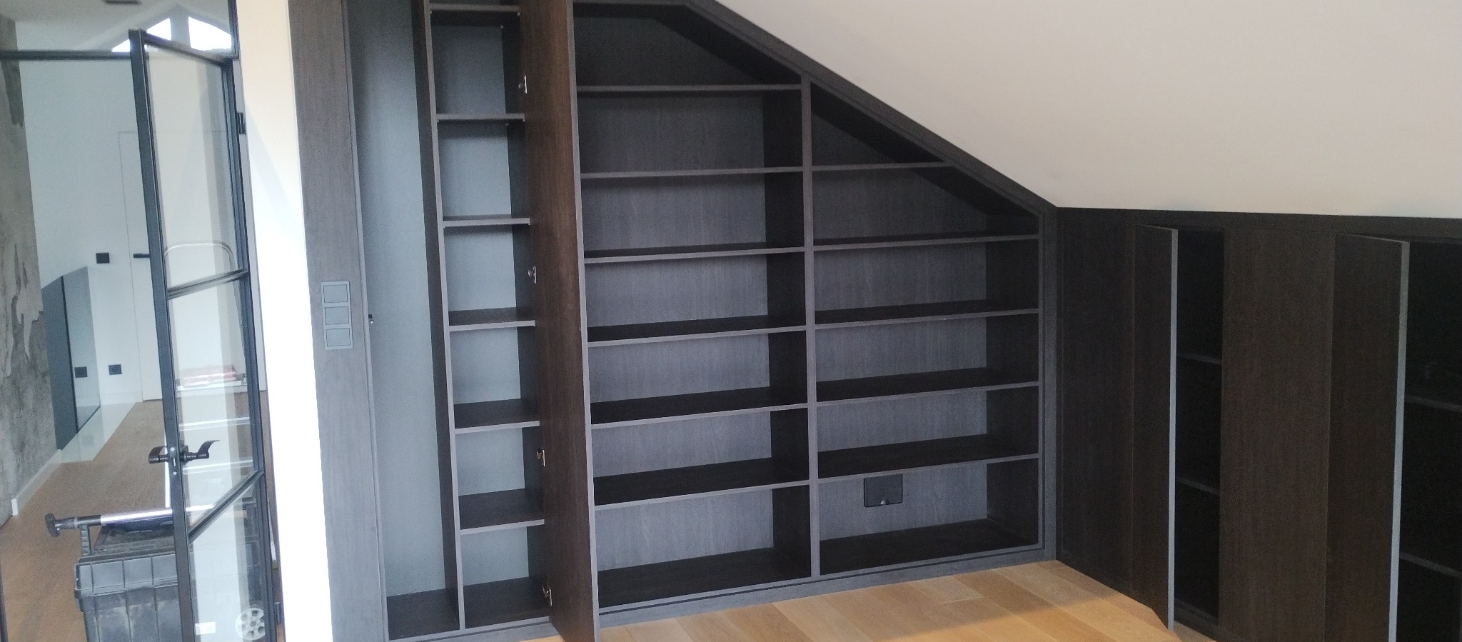 Zabudowa wnęki pod skosem z półkami, ciemne drewno, częściowo otwarte drzwiczki, widok na jasny parkiet i przeszklone drzwi.