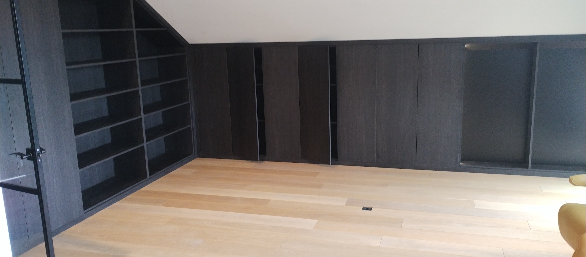 Ciemnoszara zabudowa meblowa pod skosem, z szafkami z przesuwanymi drzwiami i otwartymi półkami, na jasnej drewnianej podłodze.