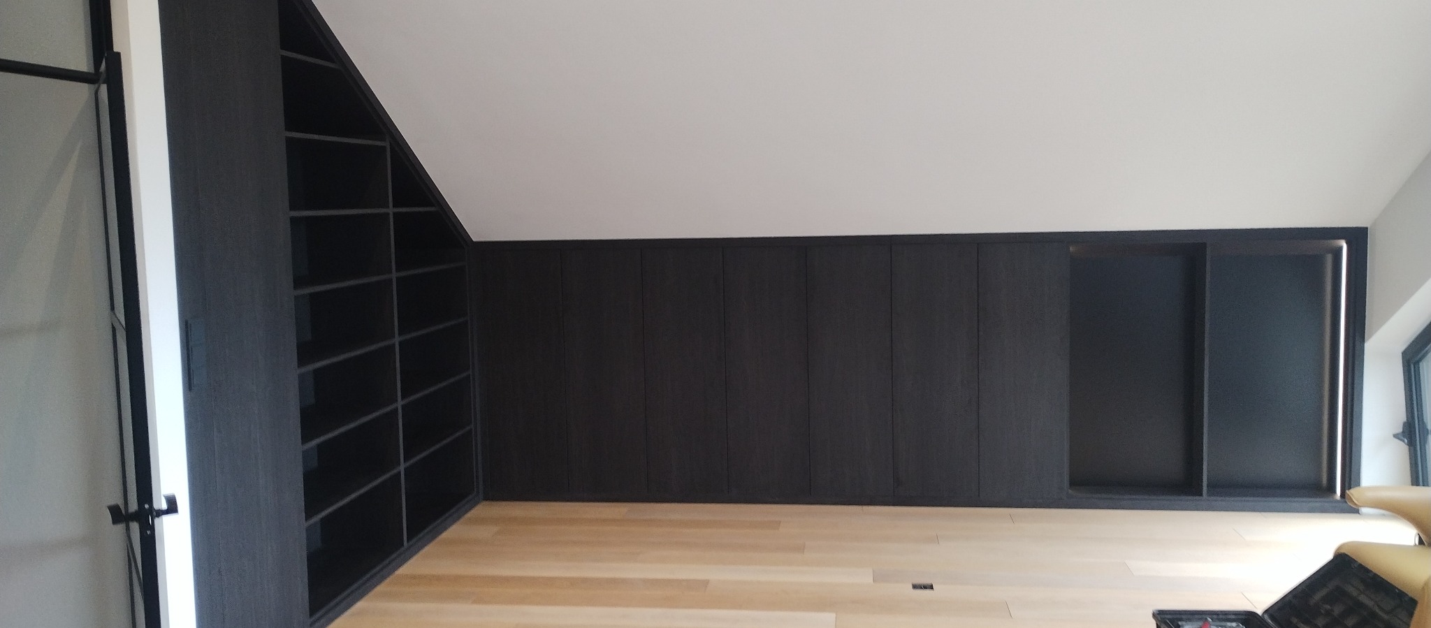 Ciemna zabudowa meblowa na wymiar w pokoju na poddaszu, z otwartym regałem na ścianie szczytowej i szafą przesuwną z podświetleniem LED.