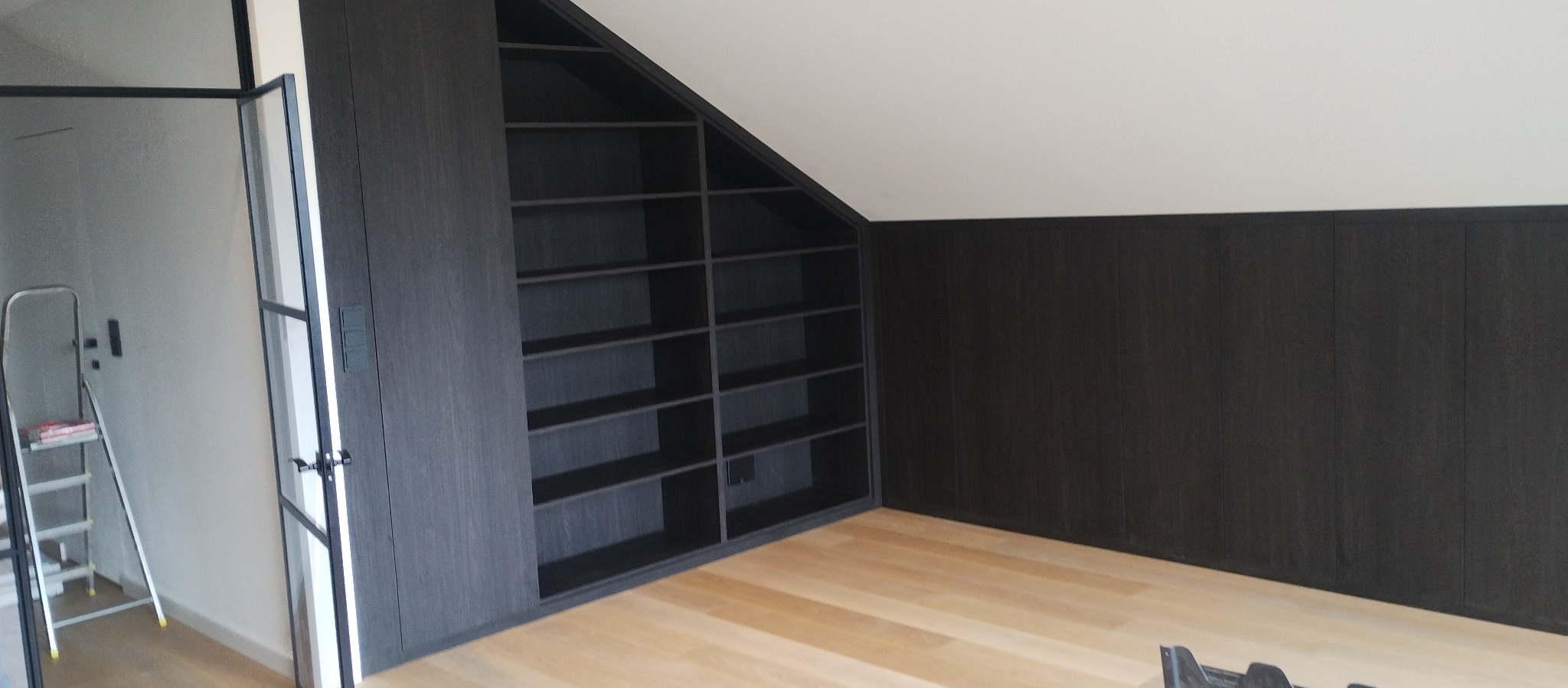 Ciemna, drewniana szafa wnękowa z półkami, dopasowana do skosu dachu, z drabiną opartą o ścianę w tle.