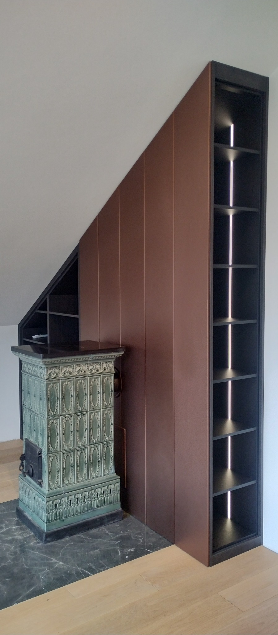 Zabudowa wnęki na poddaszu z brązowymi, gładkimi frontami szafy, obok stoi piec kaflowy z zielonymi kaflami i czarnym blatem, a po prawej stronie otwarta, czarna półka z oświetleniem LED.