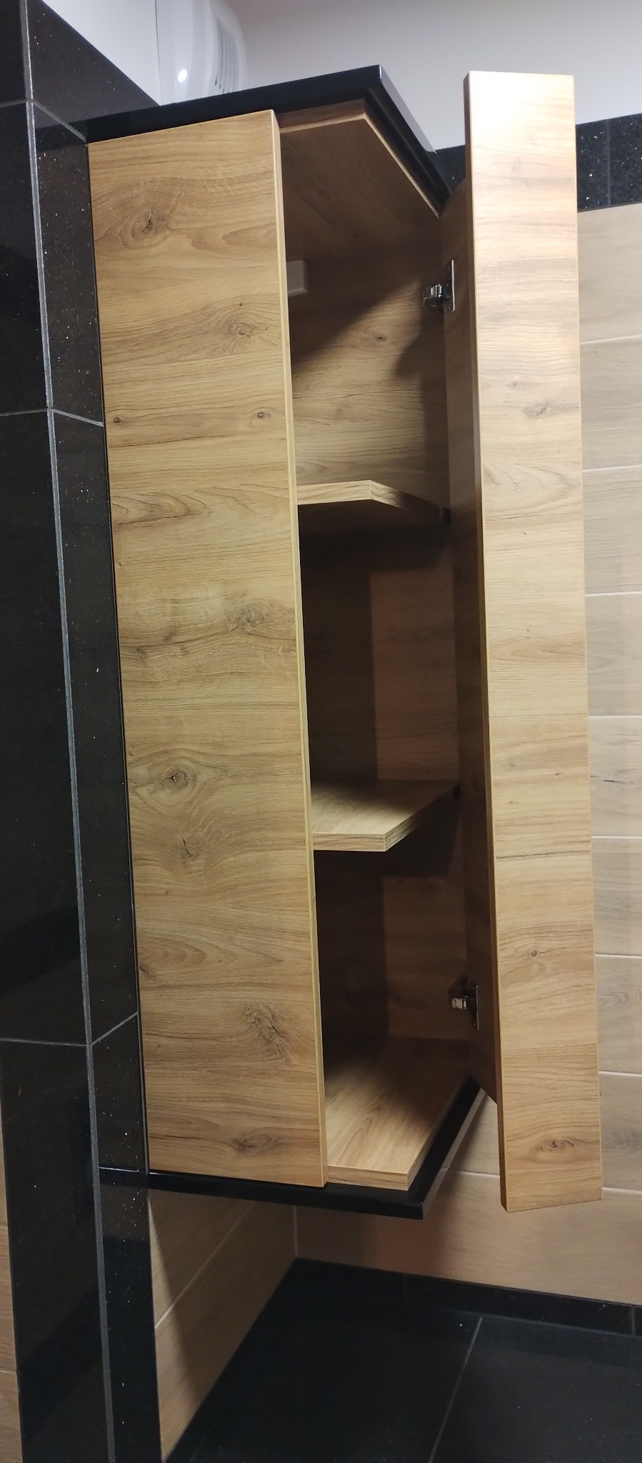 Otwarta szafka wisząca z drewnopodobnymi frontami i czarnym blatem, widoczne dwie półki wewnętrzne i metalowe zawiasy.