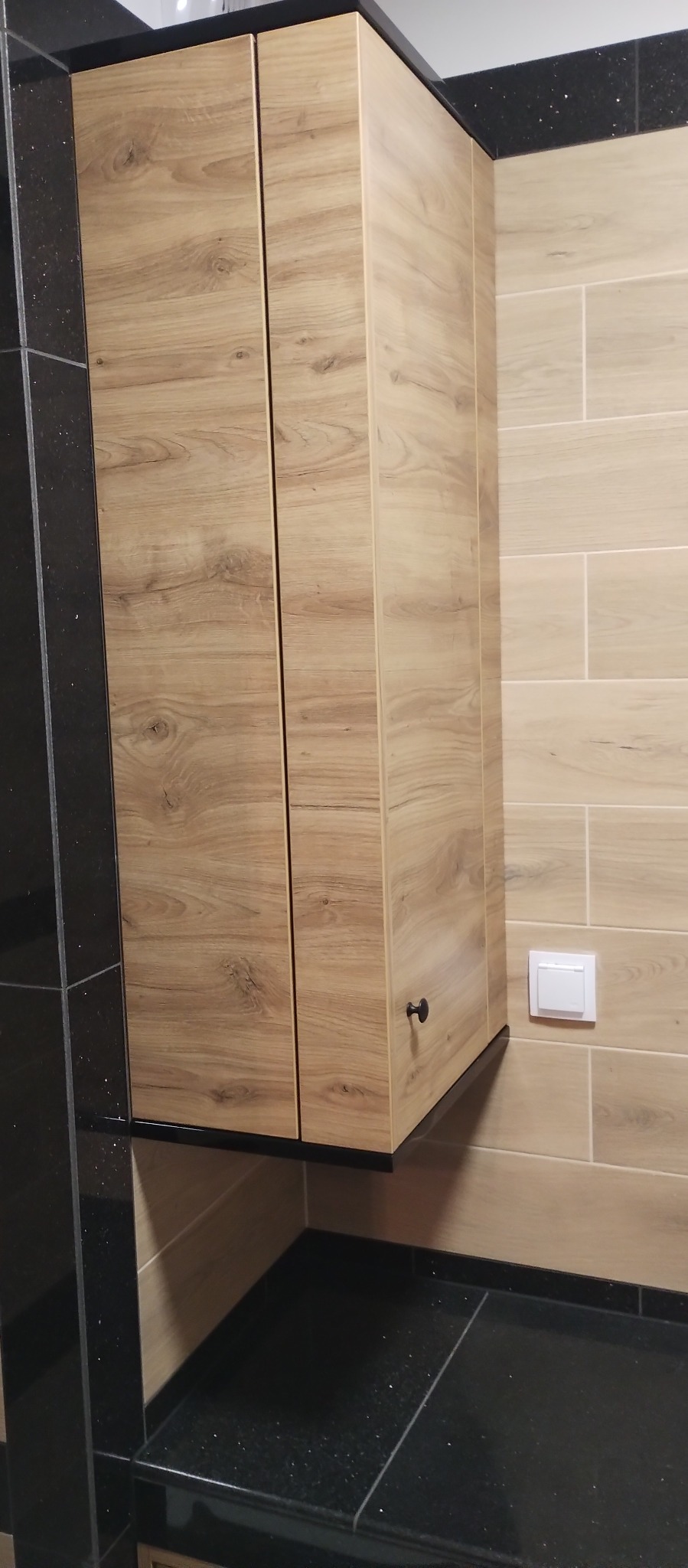 Kuchenna szafka wisząca z frontami w kolorze jasnego drewna, zamontowana nad czarnym, błyszczącym blatem, z widocznym włącznikiem światła na ścianie z płytek.