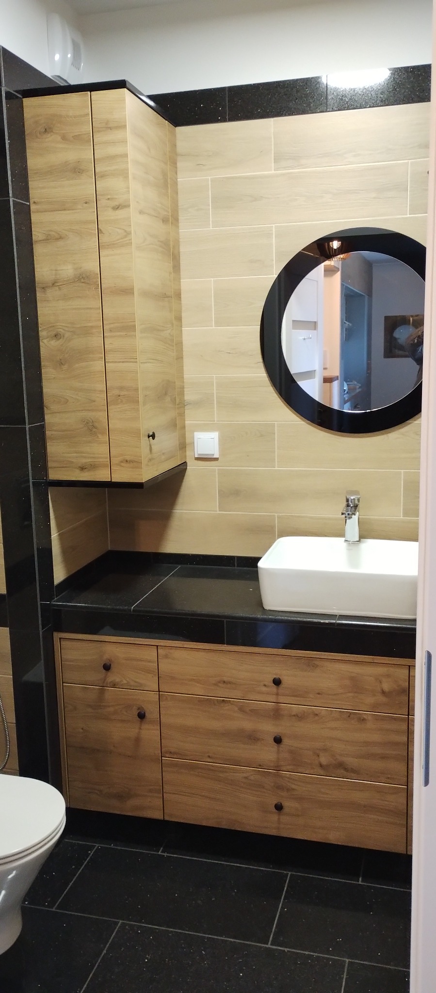 Nowoczesna łazienka z drewnianymi szafkami, czarnym blatem i okrągłym lustrem z czarną ramą, beżowe płytki na ścianie, czarna podłoga z połyskiem.
