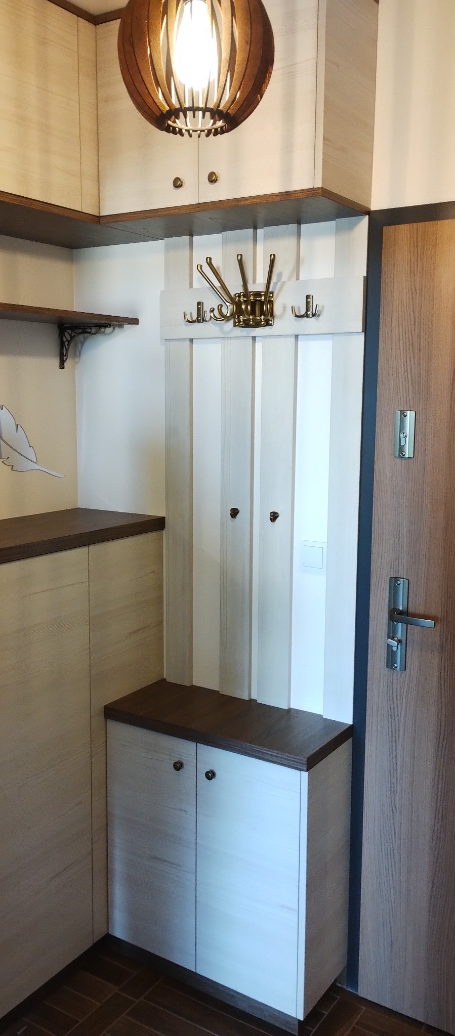 Przedpokój z zabudową na wymiar: szafka z dwiema drzwiczkami w kolorze jasnego drewna z ciemnobrązowym blatem, biały panel z wieszakami w stylu retro i okrągła lampa sufitowa z drewnianymi elementami.