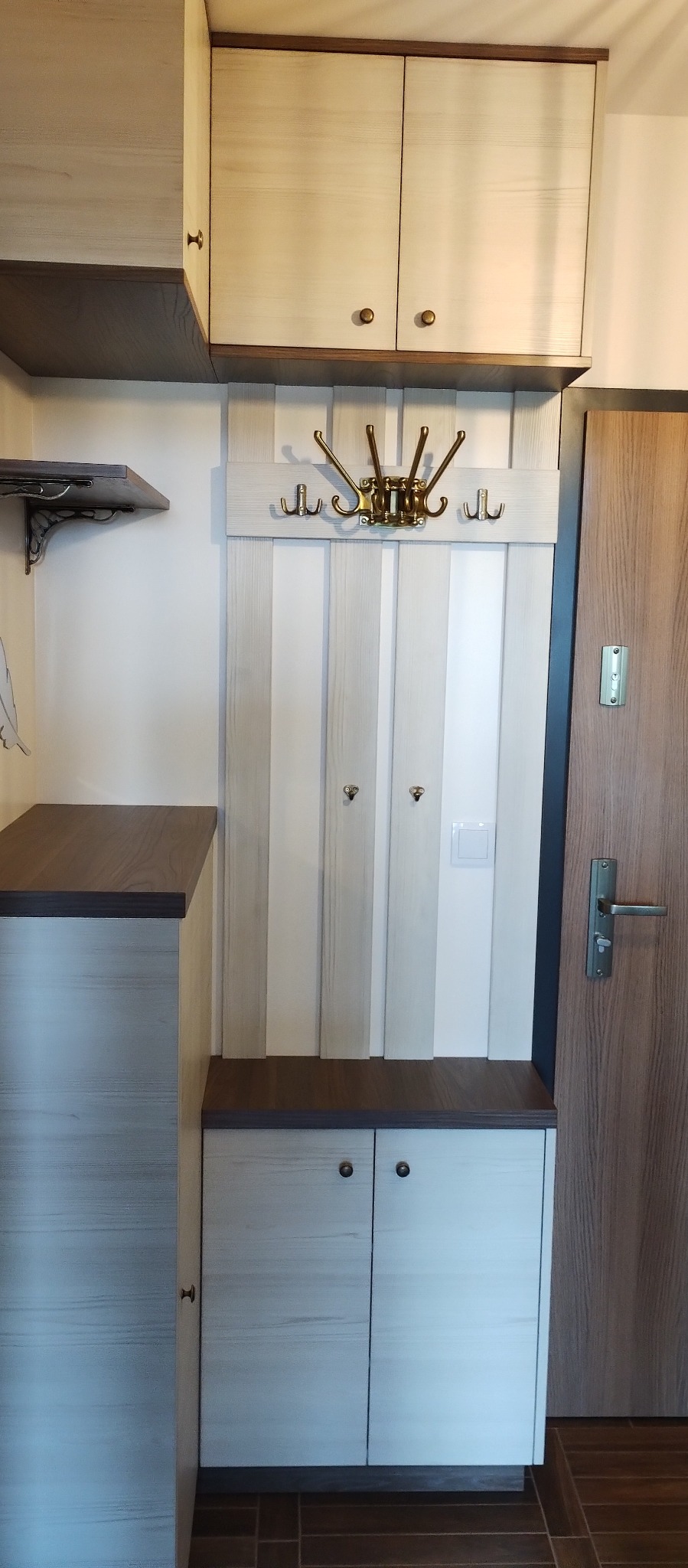 Jasna szafa w przedpokoju z drewnianymi akcentami, wbudowana wnęka z wieszakami i szafką na buty, brązowe uchwyty, drzwi wejściowe po prawej stronie.