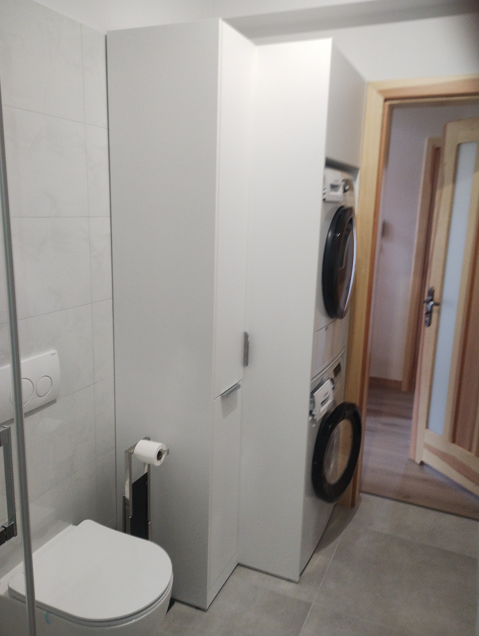 Biała szafa na wymiar w łazience obok toalety i pralko-suszarki w słabym oświetleniu.