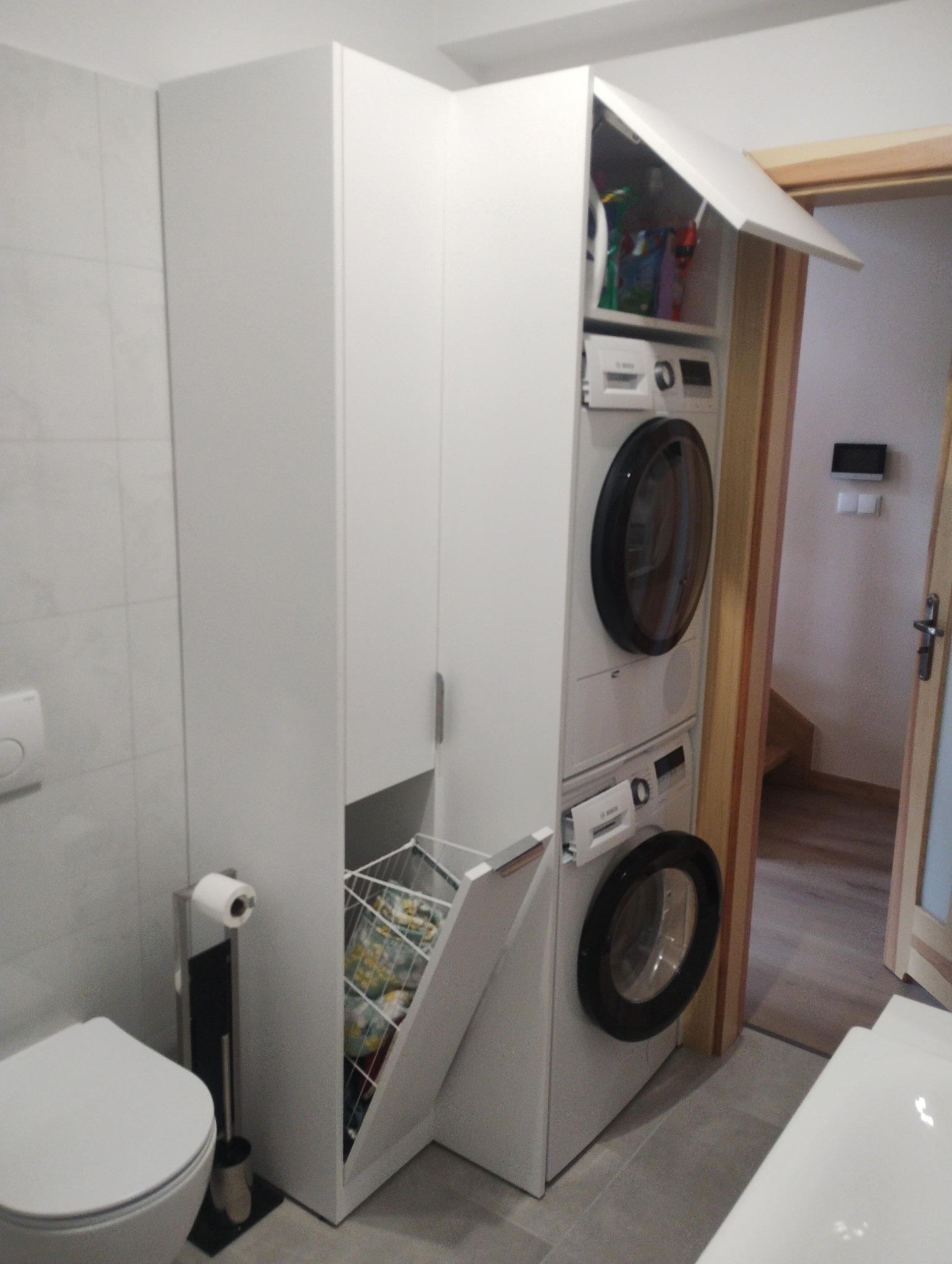 Biała zabudowa w łazience z pralką i suszarką w kolumnie, otwarty kosz na pranie z kolorowymi ubraniami, widoczna toaleta i uchwyt na papier toaletowy.