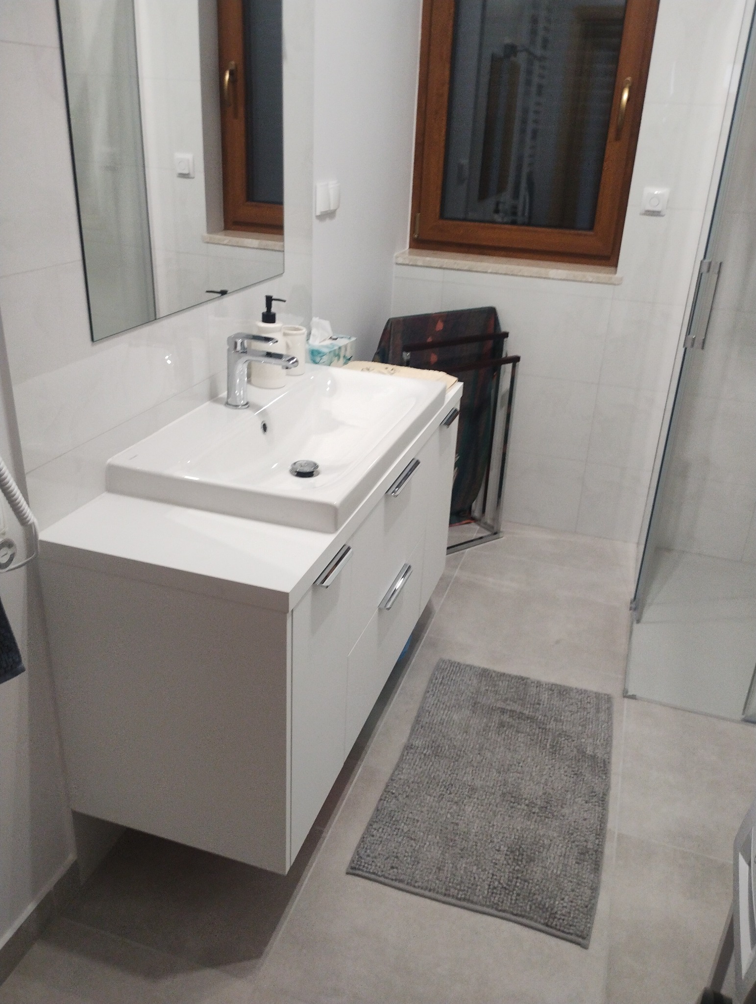 Nowoczesna biała szafka łazienkowa z umywalką nablatową i chromowaną armaturą, szary dywanik, widok na okno z drewnianą ramą i kabinę prysznicową.