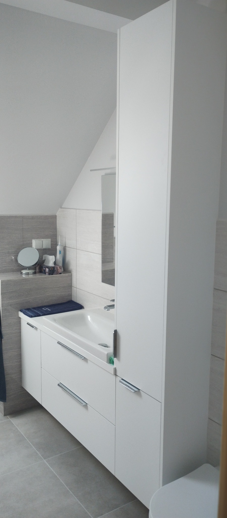 Biała szafka łazienkowa z umywalką i wysoką szafką w nowoczesnej łazience z szarymi płytkami, widoczna część toalety.
