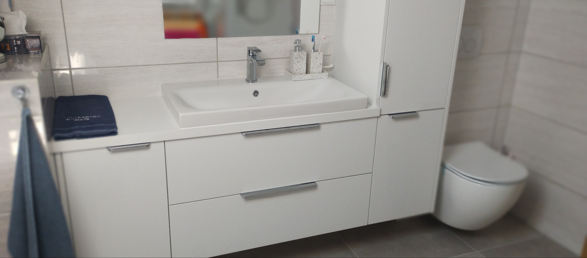 Nowoczesna, biała szafka łazienkowa z umywalką i dwoma szufladami, z wiszącą szafką boczną i toaletą w tle na tle jasnych płytek.