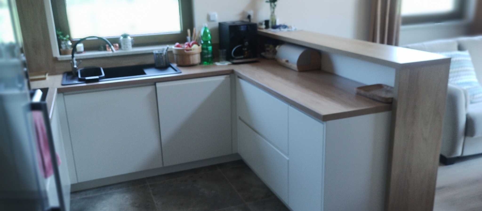 Jasna kuchnia w kształcie litery L z drewnianym blatem i barkiem, widok na okno z ziołami i akcesoriami kuchennymi.