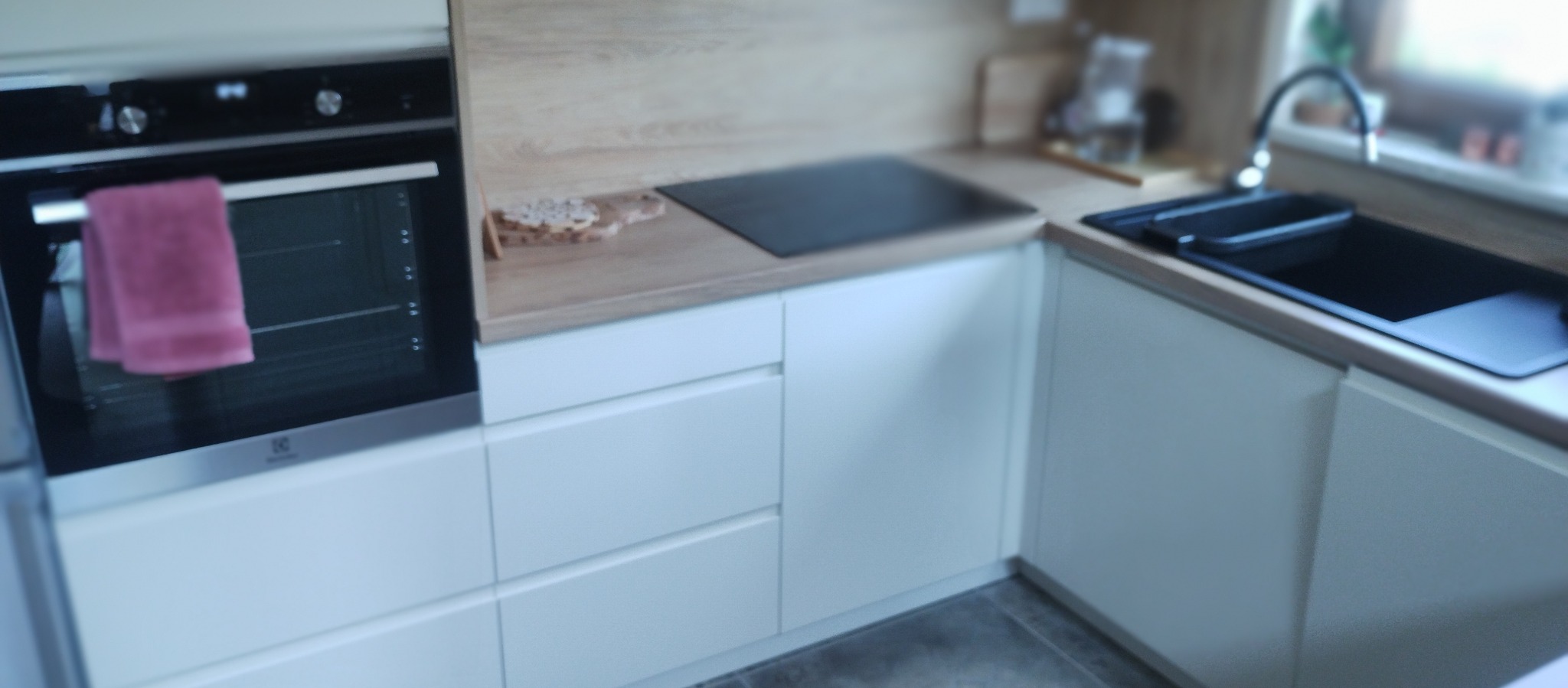 Fragment białej, nowoczesnej kuchni z drewnianym blatem, czarną płytą indukcyjną i zlewem. Na piekarniku zawieszony różowy ręcznik.